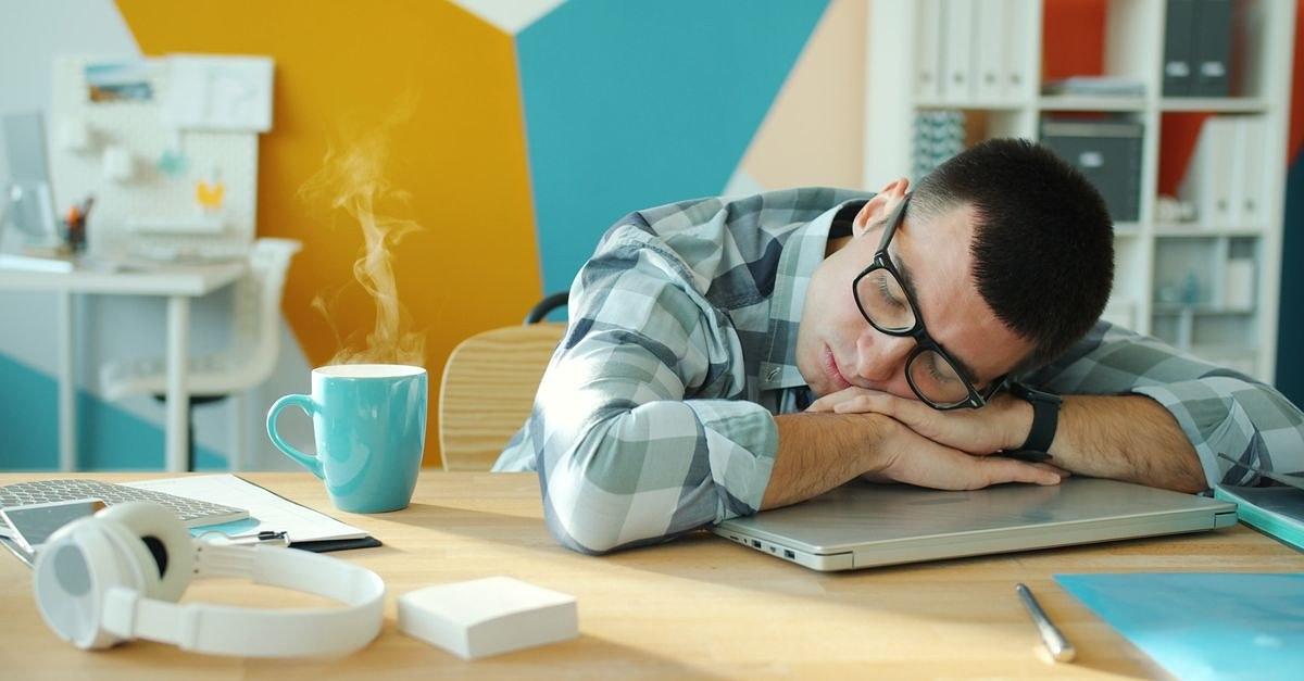 Young man sleeping on a laptop at his desk with a hot beverage nearby.