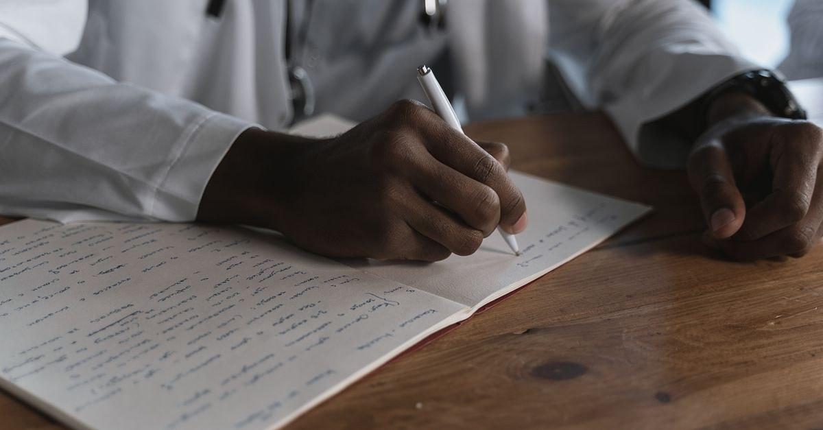 Close-up of a doctor writing notes with a pen in a medical notebook on a wooden desk.