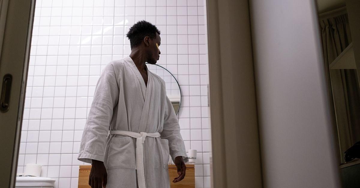 A man in a white bathrobe is looking behind him in a modern restroom with a mirror.