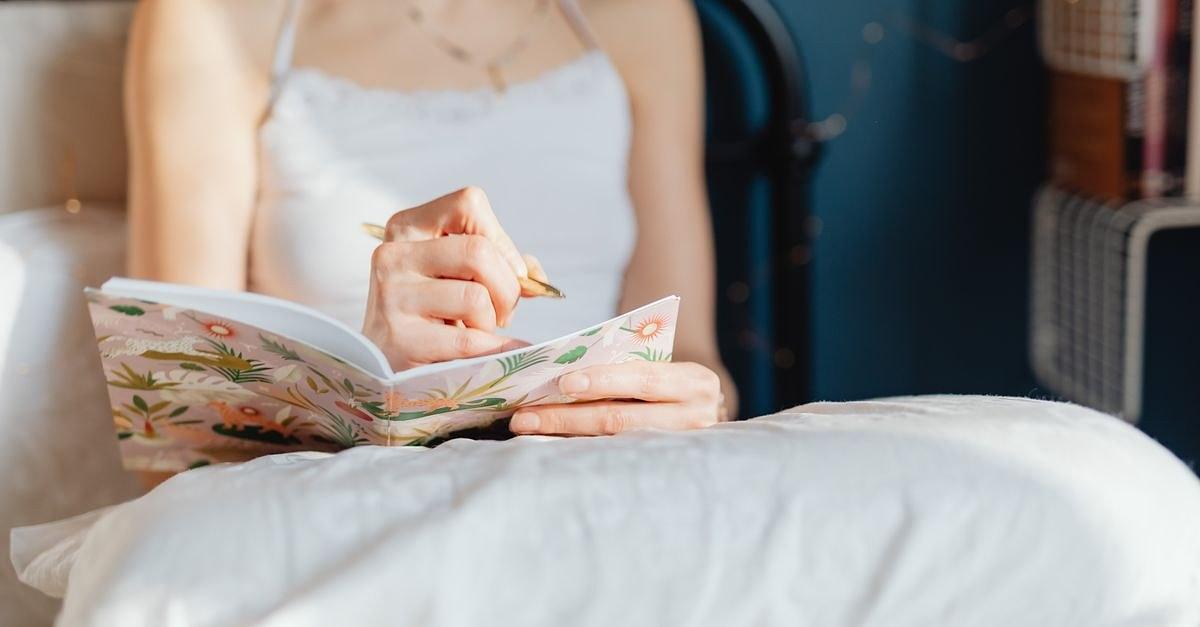 A woman in a white top writes in a decorative floral notebook while seated on her bed.