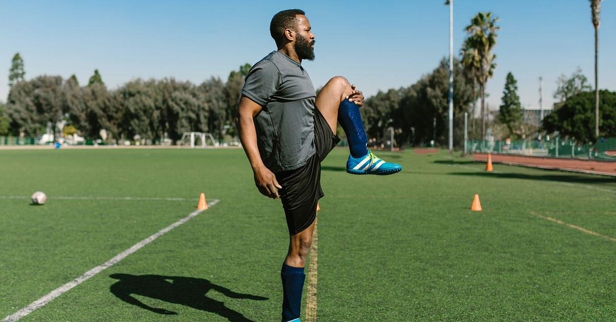 Black male athlete stretches on a green soccer field under a clear sky, preparing for training.