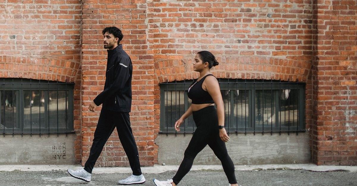 A man and woman walking briskly in sportswear against a brick wall in the city.