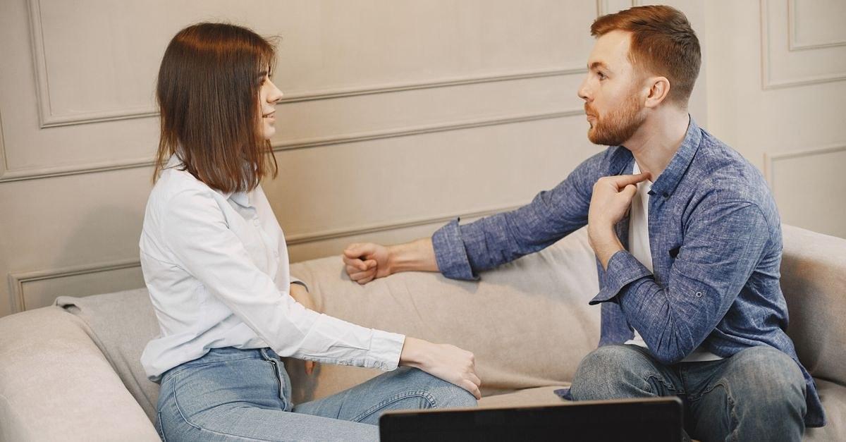 A young couple engaged in a conversation while sitting on a beige couch indoors.
