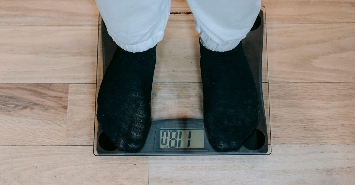 From above of crop unrecognizable plus size person in casual clothes standing on weighing scale on wooden floor