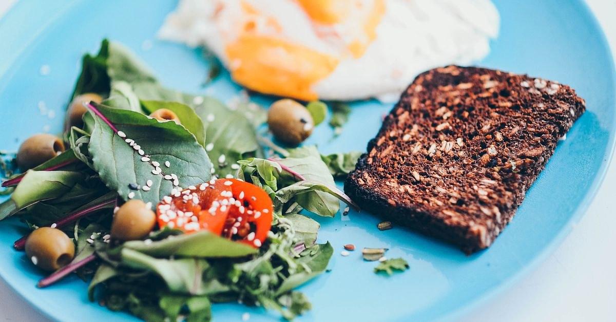 A nutritious breakfast plate featuring eggs, rye bread, salad, and olives on a vibrant blue plate.