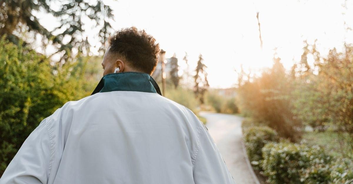 A man with headphones walking in a sunlit park, wearing a white jacket.