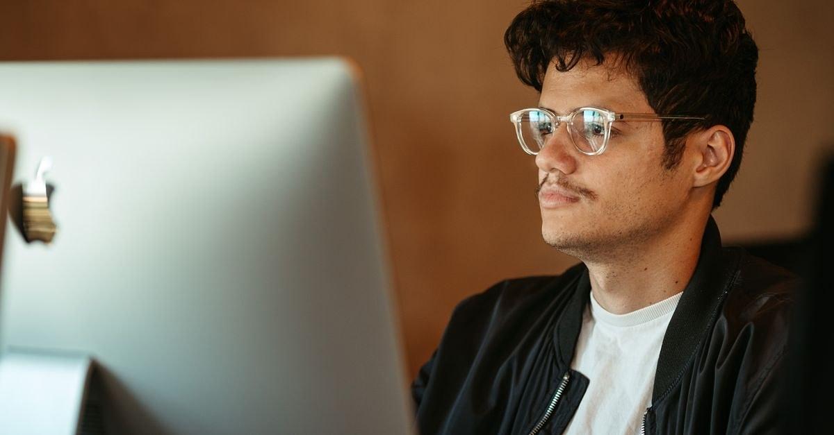 Young man in eyeglasses focusing on computer screen indoors.