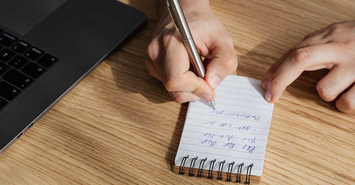 From above of crop unrecognizable man writing out information in spiral notebook while working on business plan on laptop