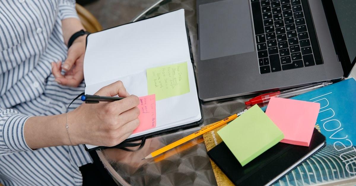 Person writing in notebook with laptop and sticky notes on a desk. Creative and organized workspace.