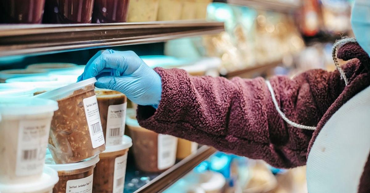 Crop anonymous female customer in protective mask reading label on frozen food in plastic container in grocery store