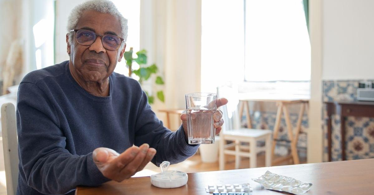 Elderly man sitting at a table at home, holding a glass of water and medication, emphasizing healthcare routine.