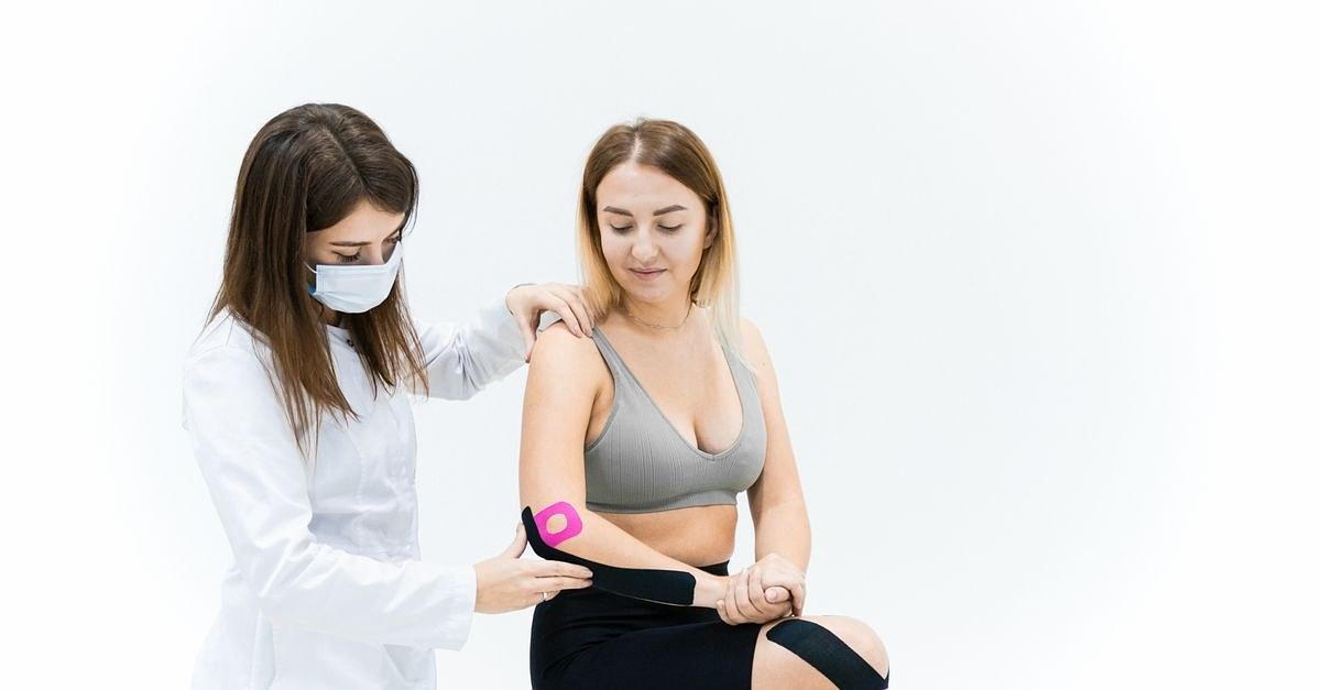 A therapist applies kinesio tape on a woman's knee during a physical therapy session on a white background.